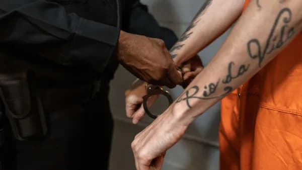 A police officer handcuffing a tattooed prisoner in an orange jumpsuit indoors.