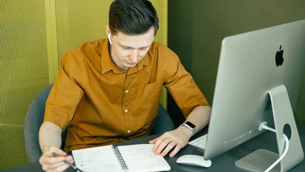 Focused young man sketching at his desk with a computer and notebook in a creative office setting.