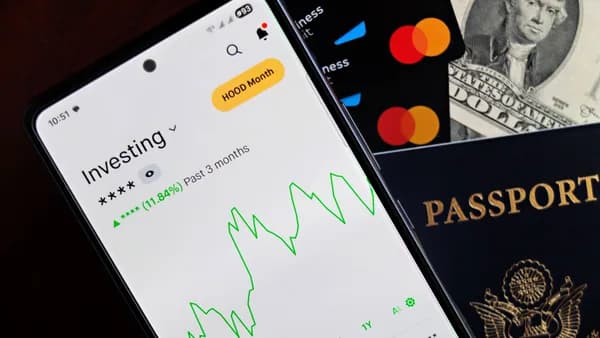 Smartphone displaying investing app, with credit cards, cash, and passport nearby, symbolizing finance