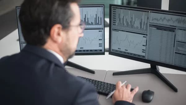 A businessman in a suit analyzing financial charts and graphs on two computer monitors indoors.