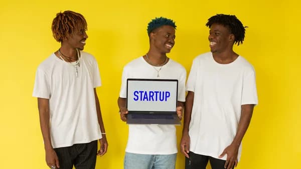 Three smiling young men in casual attire displaying a laptop that says 'Startup' on a vibrant yellow background.