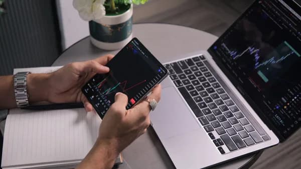 Man analyzing stock trends with laptop and mobile trading app at a modern workspace.