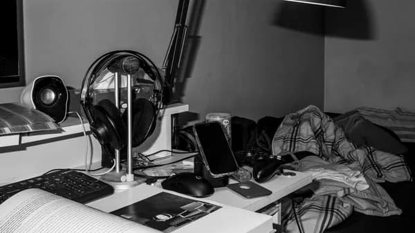 Black and white photo of a cluttered bedroom desk with tech gadgets and reading materials.