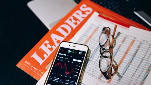 A close-up of a smartphone with cryptocurrency data next to financial reports and a magazine titled 'LEADERS'.