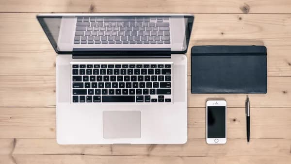 Top view of a tidy workspace featuring a laptop, smartphone, notebook, and pen on a wooden desk.