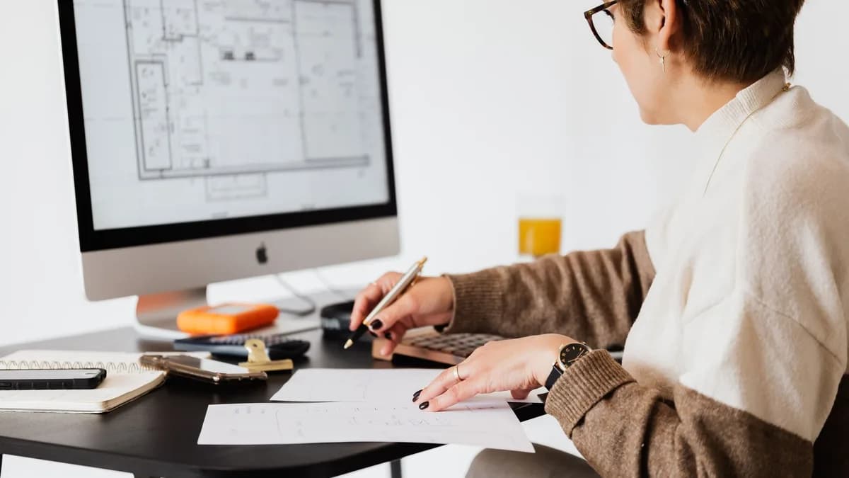 Side view of crop elegant female estate agent looking at floor plan on computer screen and noting down details on paper in light modern office