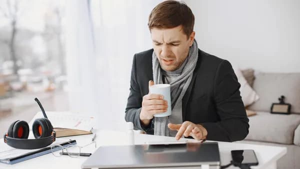 Unwell businessman working at home holding tea while sitting at a desk with a laptop.