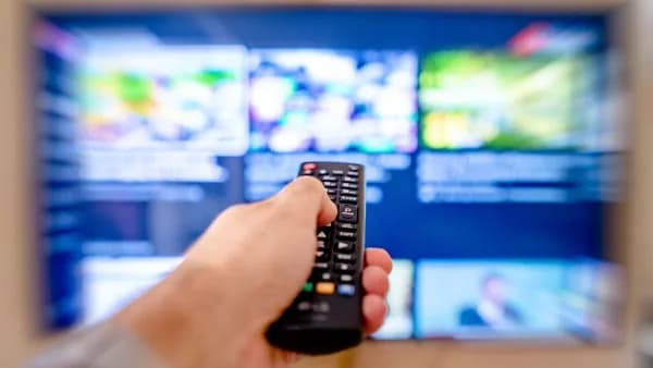 Hand holding remote control pointing at a blurred TV screen in a cozy indoor setting.