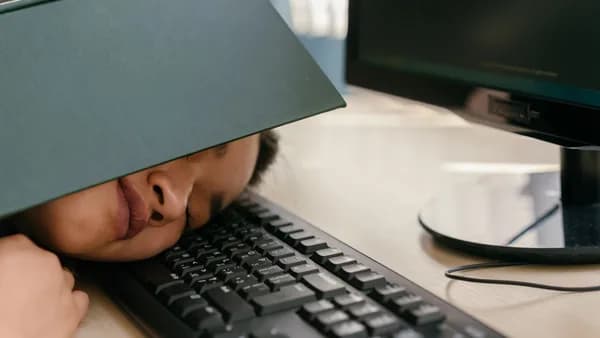 A young woman falls asleep on a keyboard with a textbook on her face, depicting study fatigue.