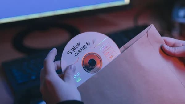 A close-up shot of hands inserting a handwritten CD into an envelope, with a computer in the background.