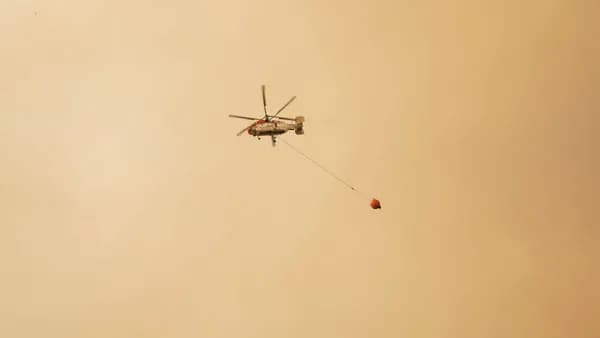 Helicopter carries water bucket against hazy sky, illustrating aerial firefighting during a wildfire.