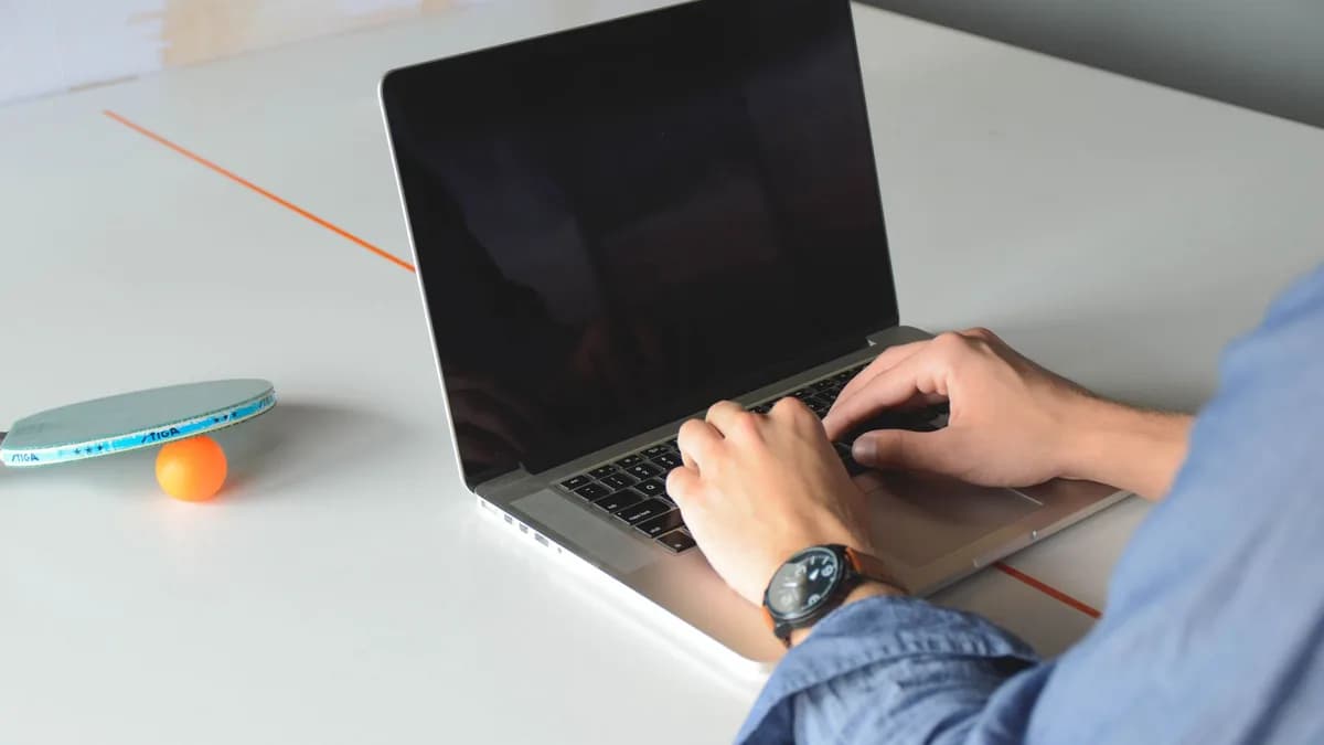A person typing on a laptop in an office, with a ping pong paddle and ball nearby.