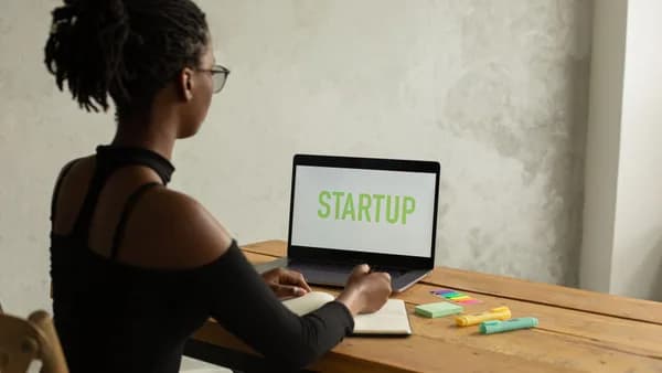 Back view of anonymous young African American businesswoman with bare shoulder and eyeglasses sitting at table with netbook with startup on screen and writing notes in copybook