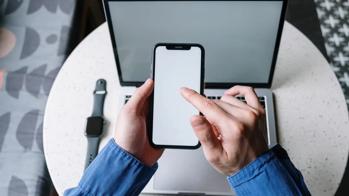 Hands operating a smartphone above a laptop with a smartwatch nearby, showcasing modern technology.