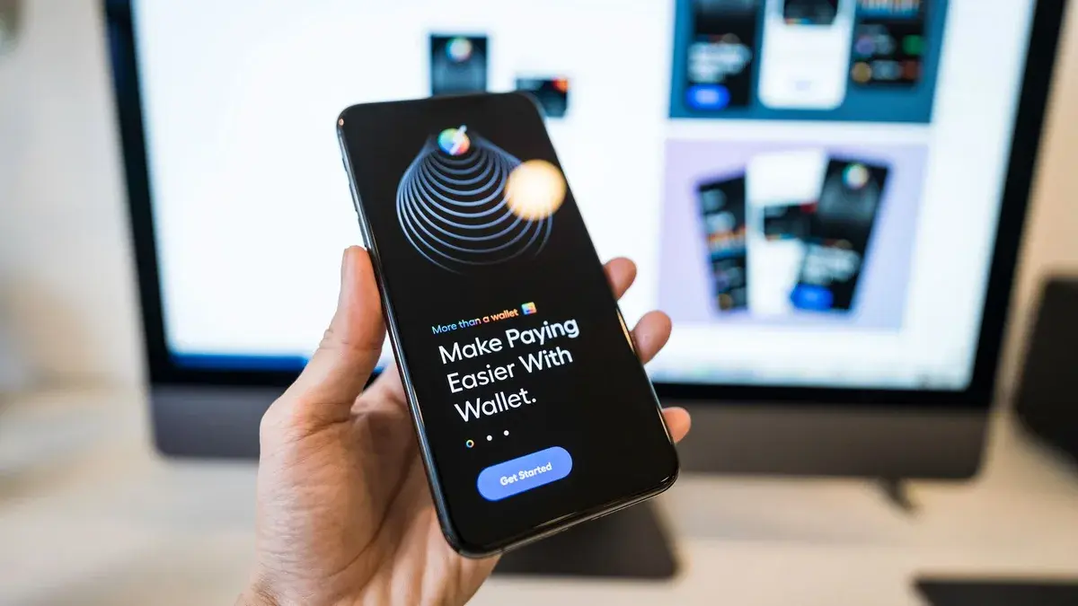Hand holding smartphone displaying digital wallet app interface, blurred monitor in background.