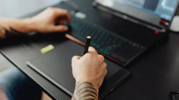 Close-up of a tattooed hand using a stylus on a digital tablet with a laptop, showcasing digital artistry.