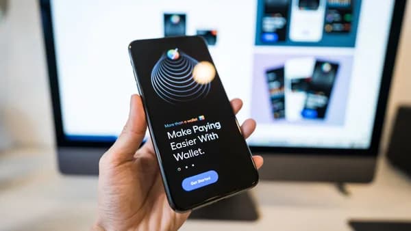 Hand holding smartphone displaying digital wallet app interface, blurred monitor in background.
