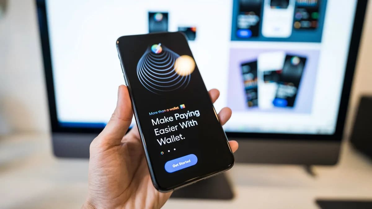 Hand holding smartphone displaying digital wallet app interface, blurred monitor in background.