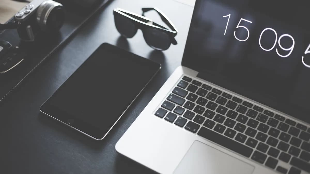 A flat lay of a modern workspace featuring a laptop, tablet, camera, and sunglasses.