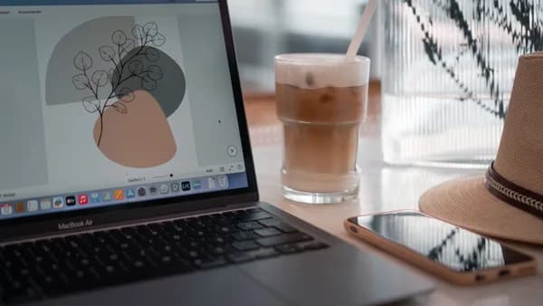 A modern workspace featuring a laptop, iced coffee, smartphone, and hat on a table.