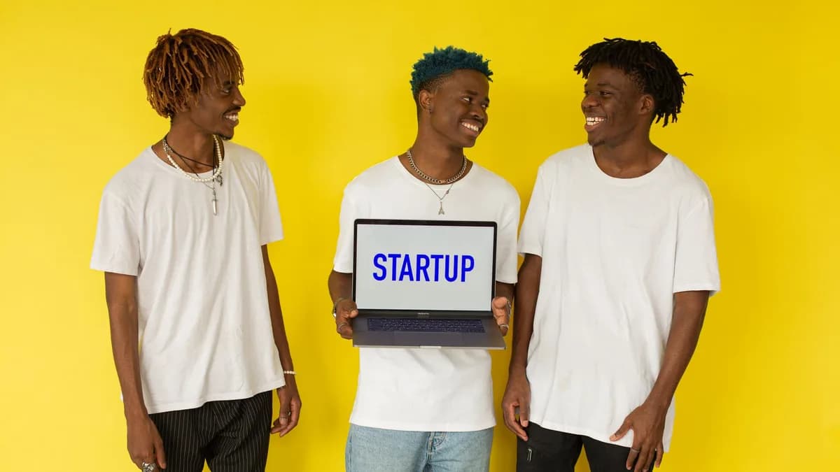 Three smiling young men in casual attire displaying a laptop that says 'Startup' on a vibrant yellow background.