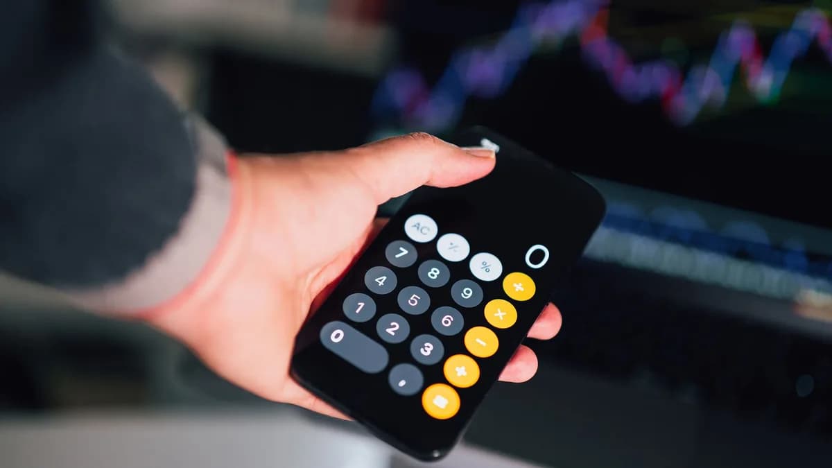 Close-up of a hand holding a smartphone calculator with financial charts on a screen in the background.