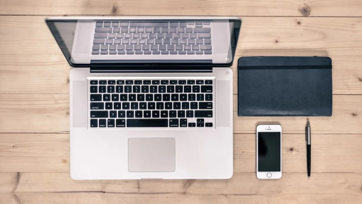 Top view of a tidy workspace featuring a laptop, smartphone, notebook, and pen on a wooden desk.