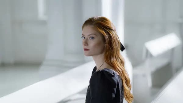 Portrait of a woman in a black dress sitting indoors, looking away thoughtfully.