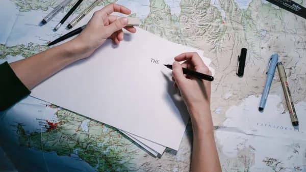 Hands writing on paper over a detailed world map with various pens.