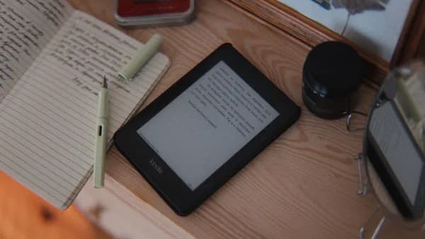 A cozy desk setup featuring an e-reader, open notebook, and pen on a wooden table.
