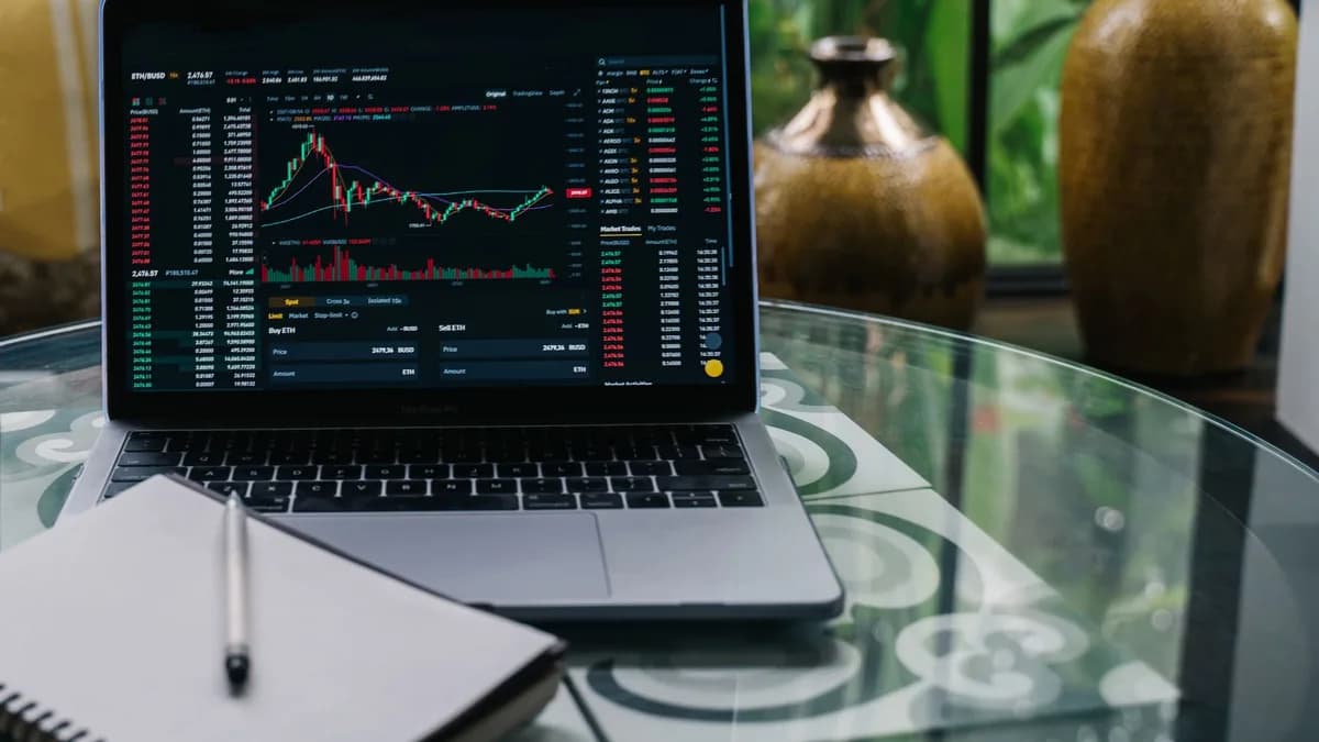 Laptop displaying cryptocurrency stocks and graphs on a glass table with a notepad.