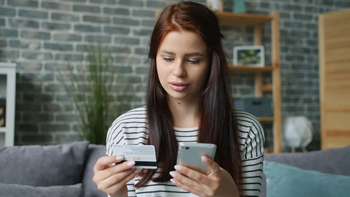 Focused woman shopping online with credit card and smartphone indoors.