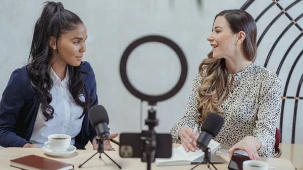 Positive blogger asking questions to African American speaker and recording interview on cellphone at table with microphones