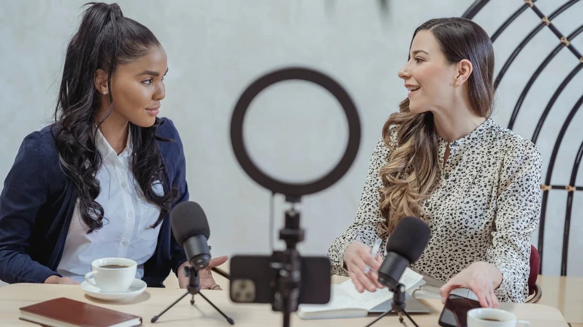 Positive blogger asking questions to African American speaker and recording interview on cellphone at table with microphones