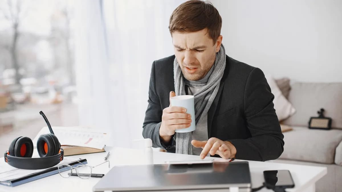 Unwell businessman working at home holding tea while sitting at a desk with a laptop.