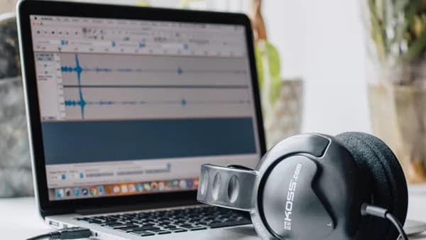 Close-up of a laptop with audio editing software and headphones in a home office setting.
