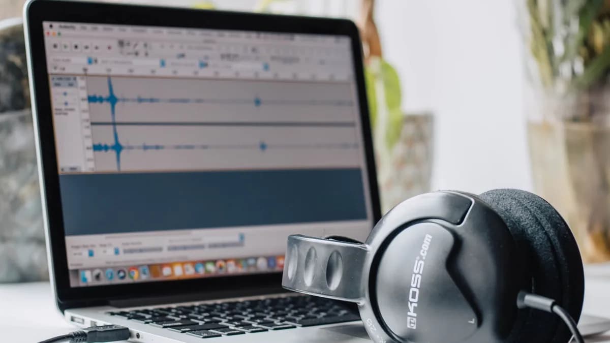 Close-up of a laptop with audio editing software and headphones in a home office setting.
