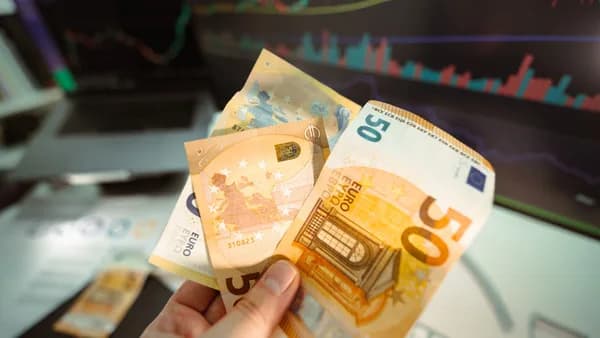 Hand holding Euro banknotes in front of a trading screen showing financial graphs.