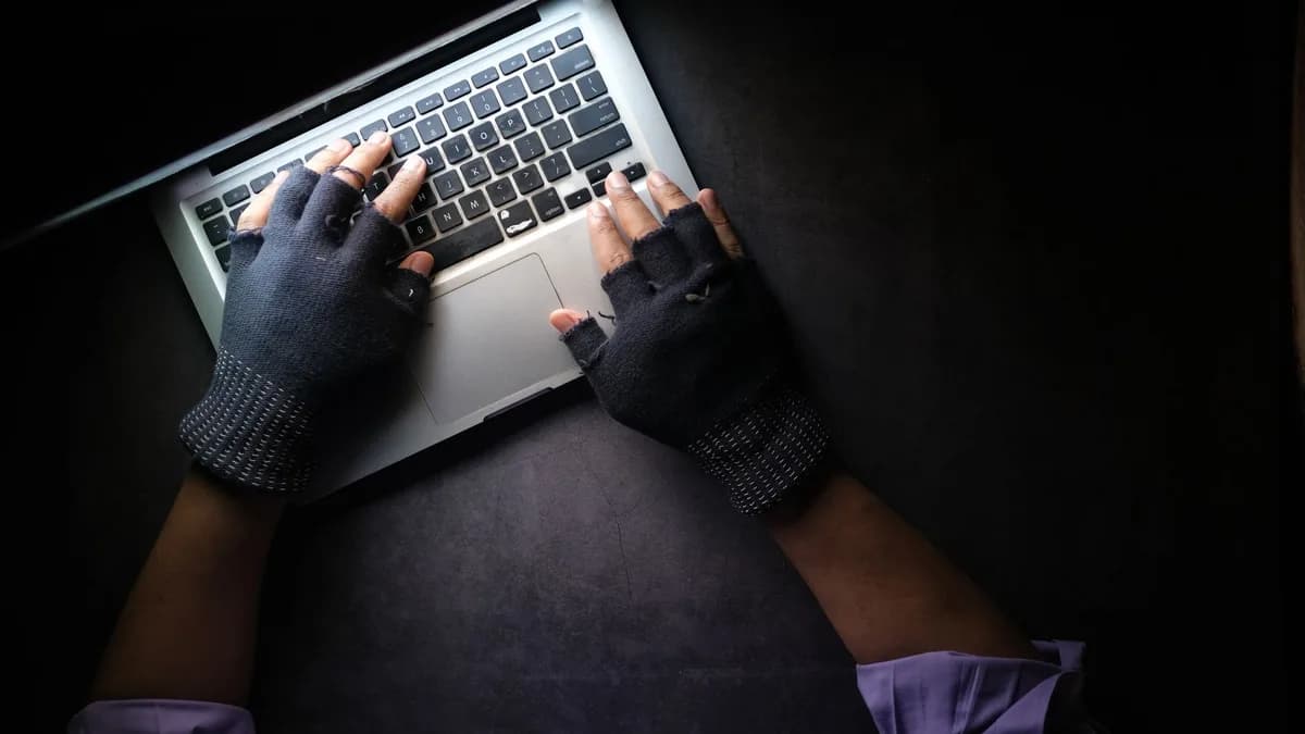 Hacker in fingerless gloves typing on laptop keyboard from above in a dark setting.