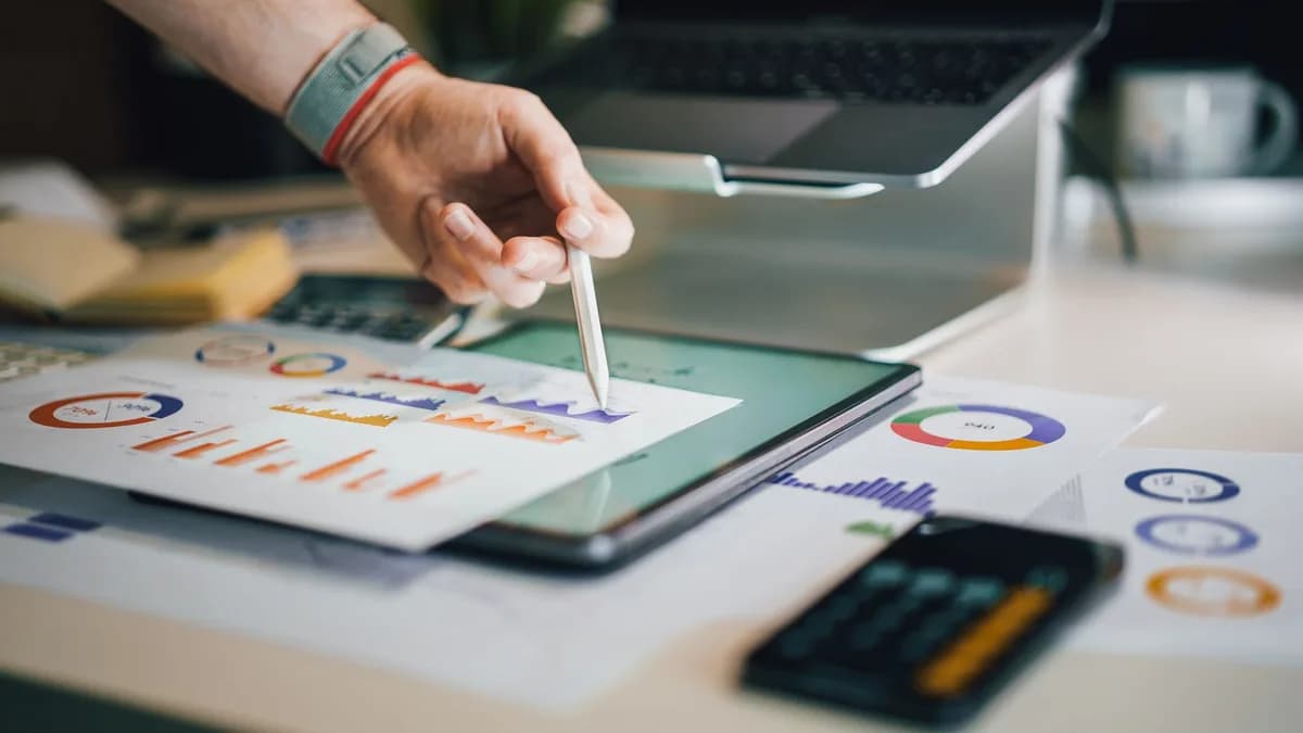 Hand using a tablet and charts on desk to analyze business data. Ideal for finance, business, and technology themes.