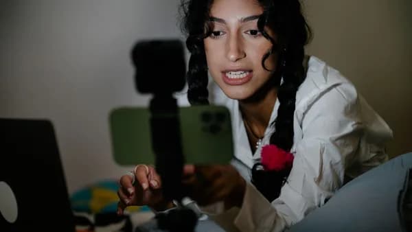 A young woman with braided hair records a video using her smartphone and laptop indoors.